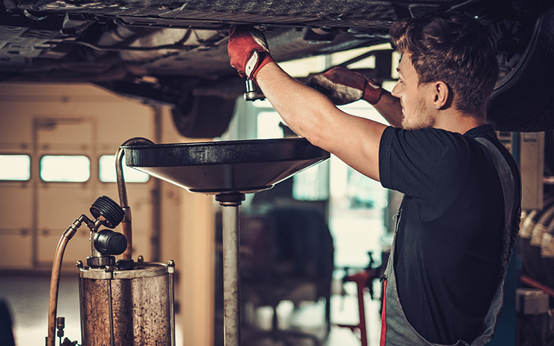 Mechanic wearing gloves performing an oil change under a lifted car in a garage.