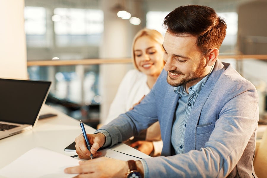 A smiling man signing a document with a happy woman beside him.