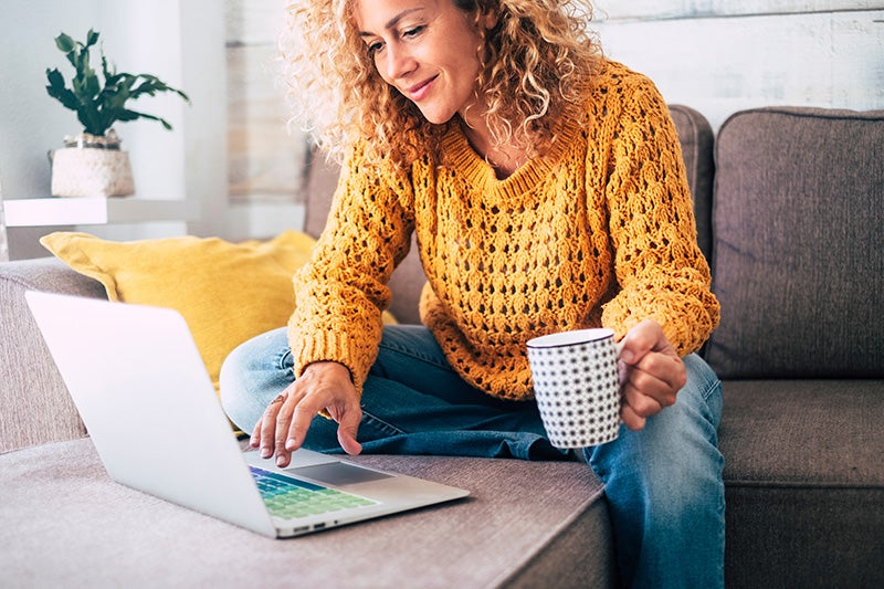 A woman sitting on a sofa, using a laptop while holding a mug of coffee.